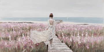 Een vrouw in een witte jurk wandelt over een houten pad door een veld vol roze en paarse bloemen, met de zee op de achtergrond.