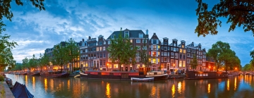 Pittoreske Amsterdamse grachten met historische gebouwen, groene bomen en reflecties van de avondverlichting op het water.