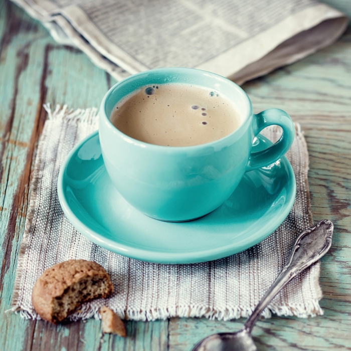 Turquoise kopje met koffie op een wit linnen servetje, omringd door een koekje en een zilveren lepel.
